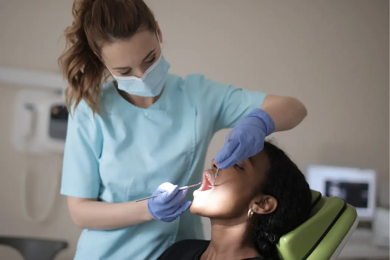 A dentist in a mask examines a patient's mouth in a dental office using dental tools.