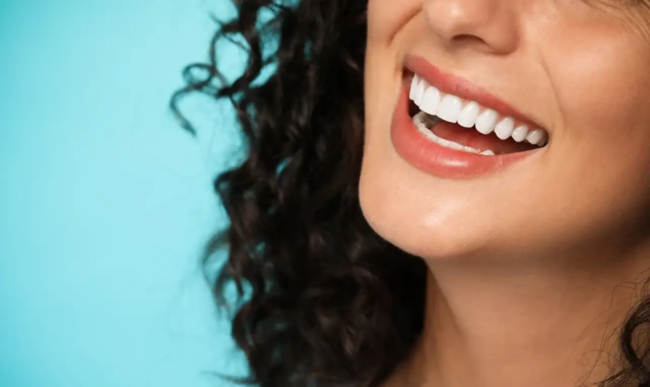 A woman with curly hair smiling, showing white teeth, against a light blue background.