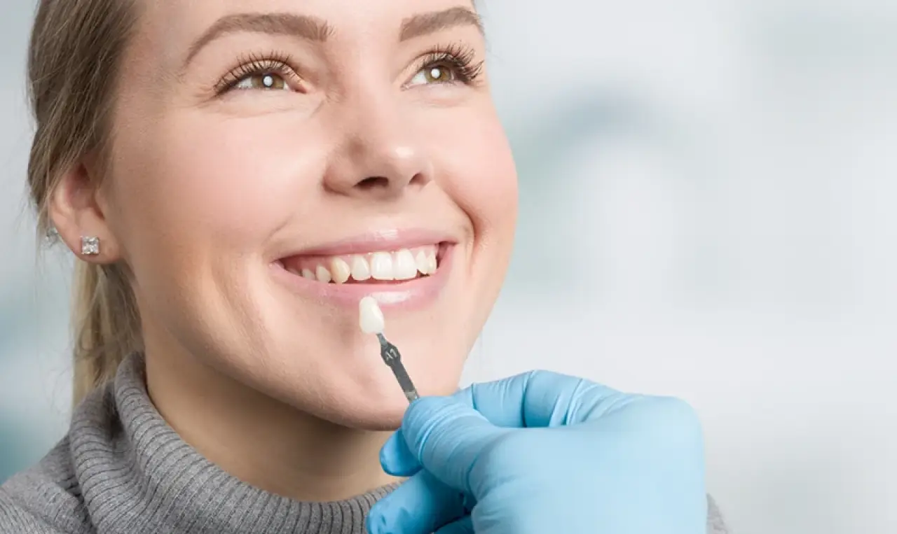 A woman smiles as a gloved hand holds a dental veneer to her teeth for comparison.