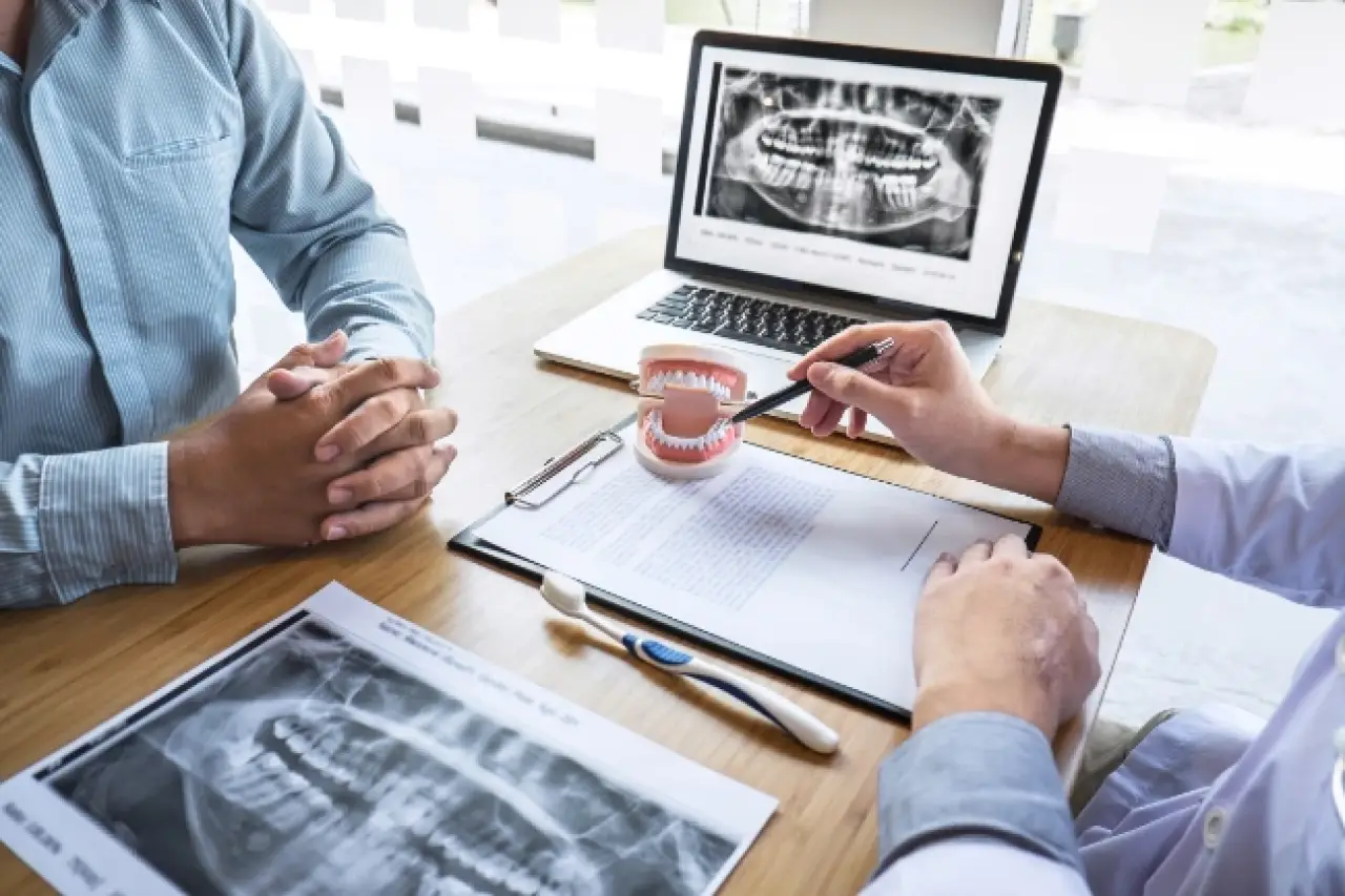 Two people discuss dental X-rays with a model of teeth, a laptop, and toothbrush on a desk.