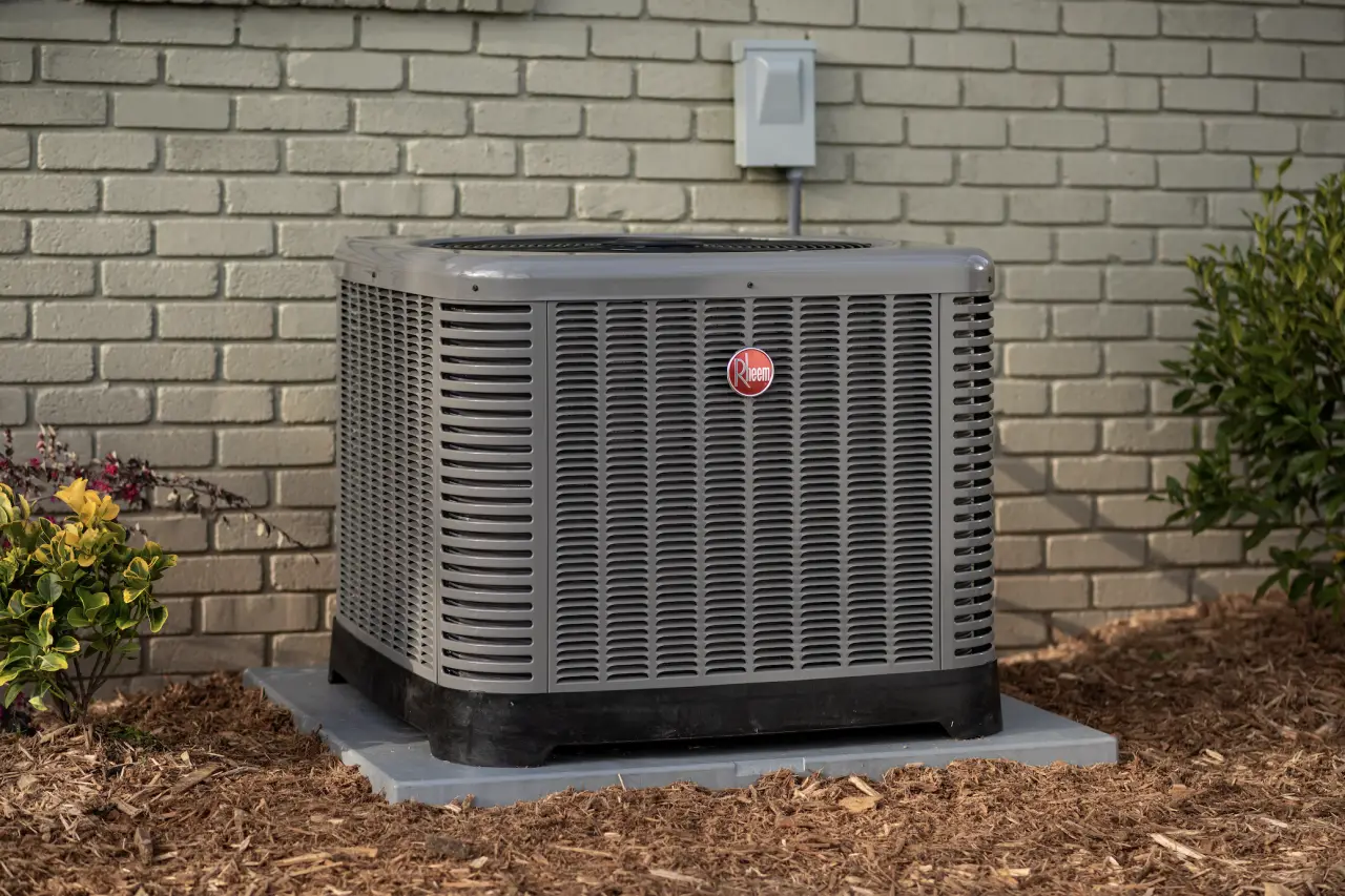 A gray outdoor air conditioning unit sits on a concrete pad next to a brick wall and plants.