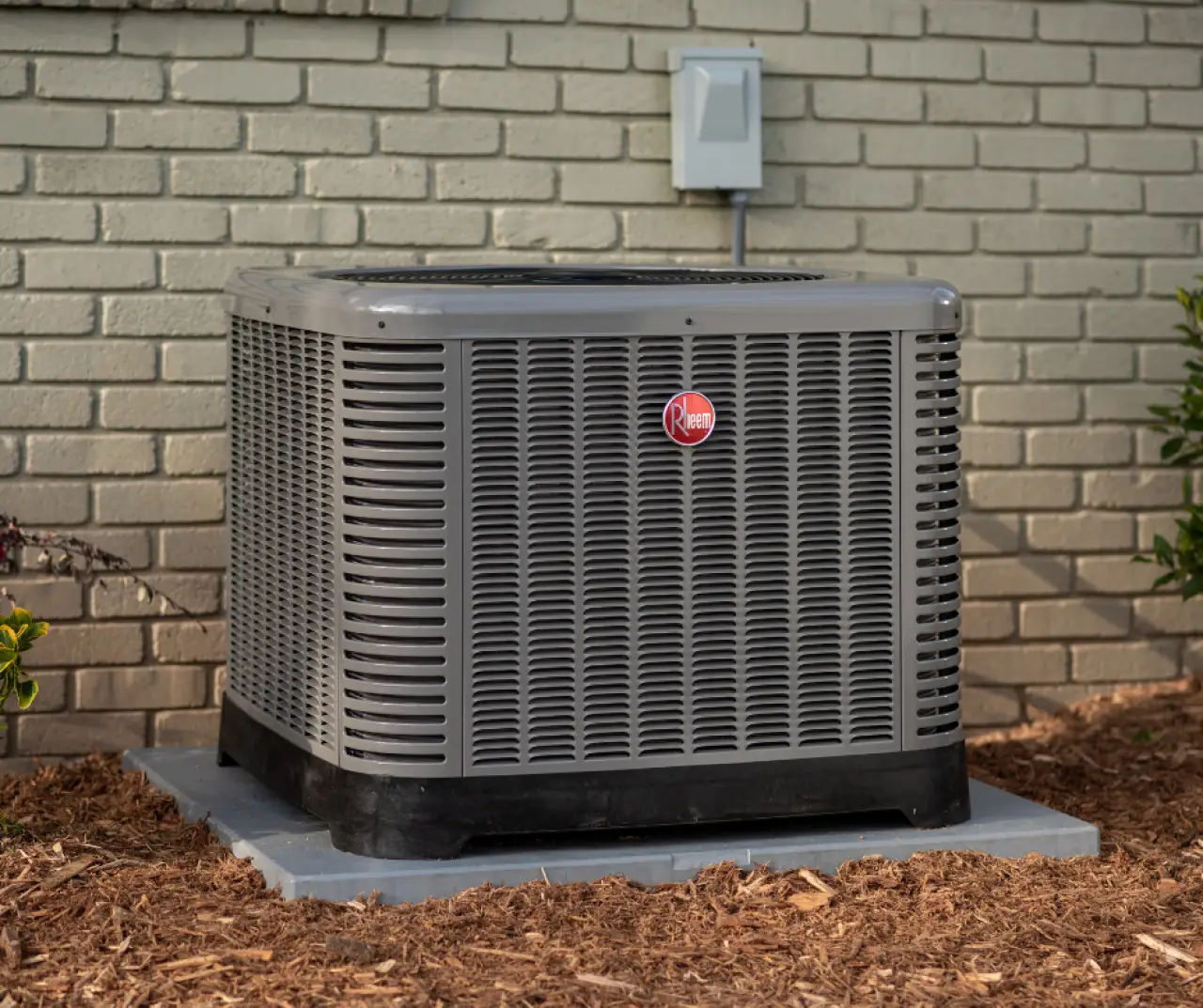 A gray outdoor air conditioning unit sits on a concrete pad next to a brick wall.