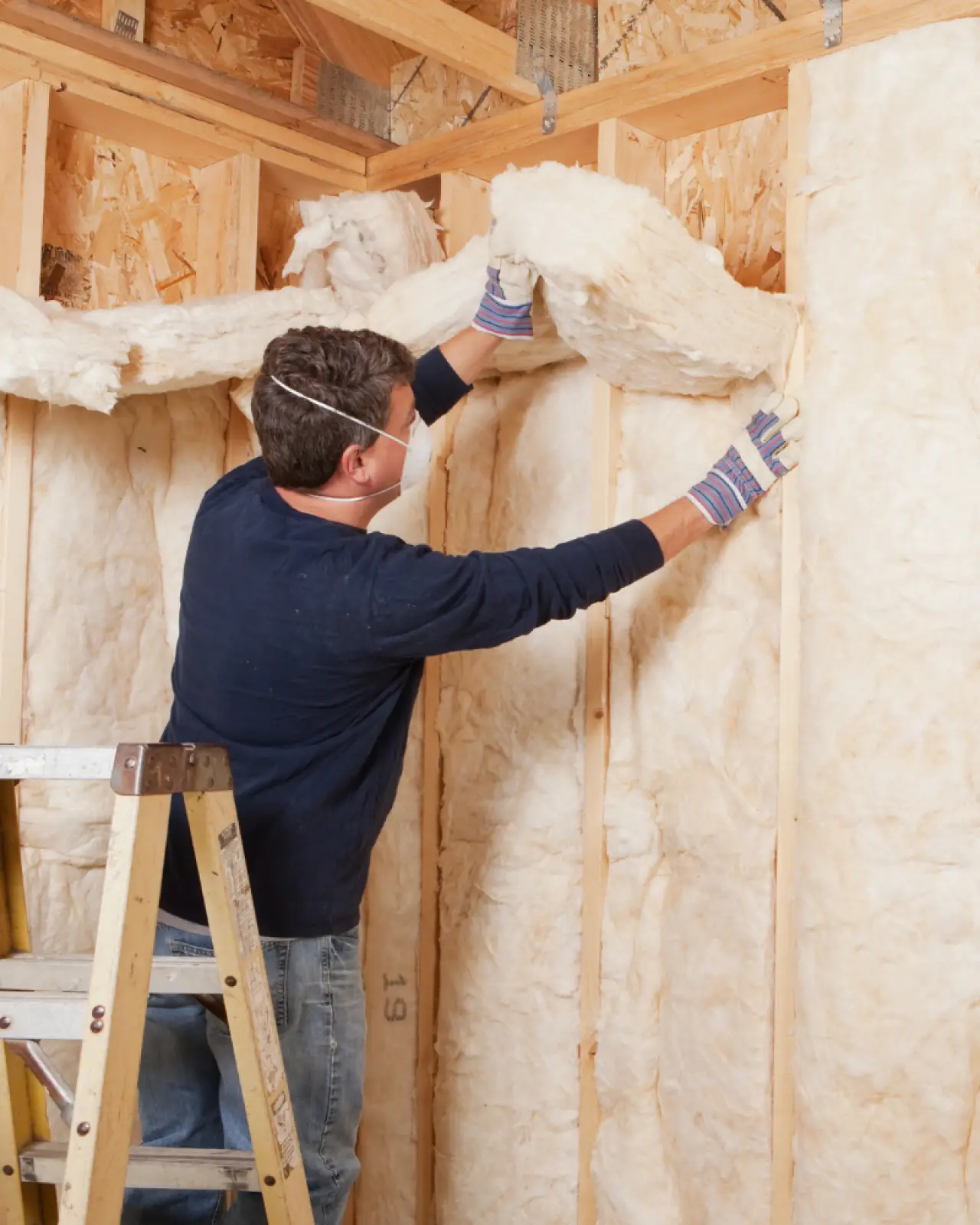 A worker wearing a respirator installs fiberglass insulation between wooden wall studs.