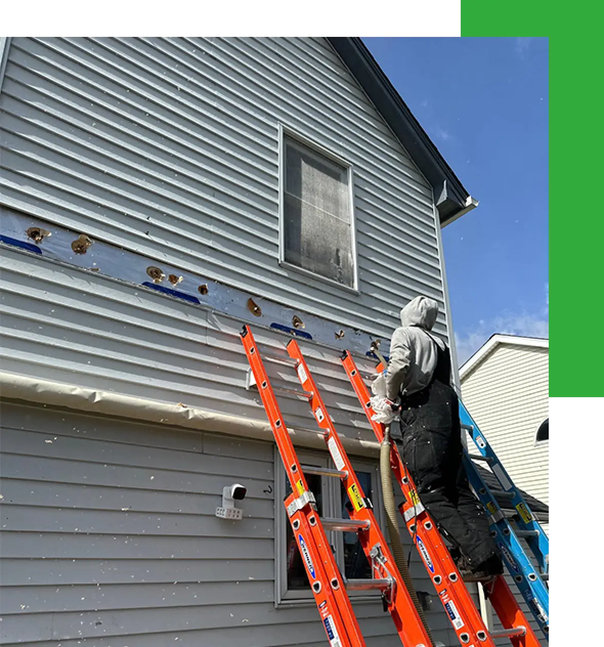 Person on ladder repairing siding on a two-story house under a blue sky.