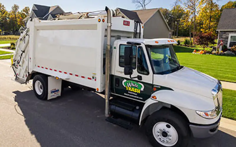 White garbage truck parked on suburban street with houses and green lawns in the background.