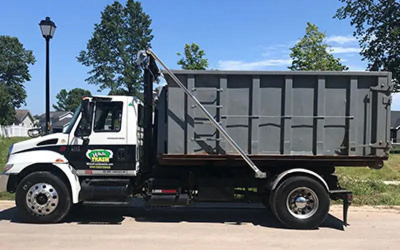 White dump truck with a large gray container parked on a sunny street with trees and grass.