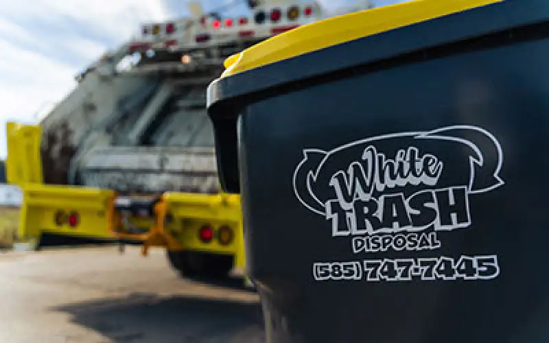 A trash bin labeled White Trash Disposal near a garbage truck on a sunny day.