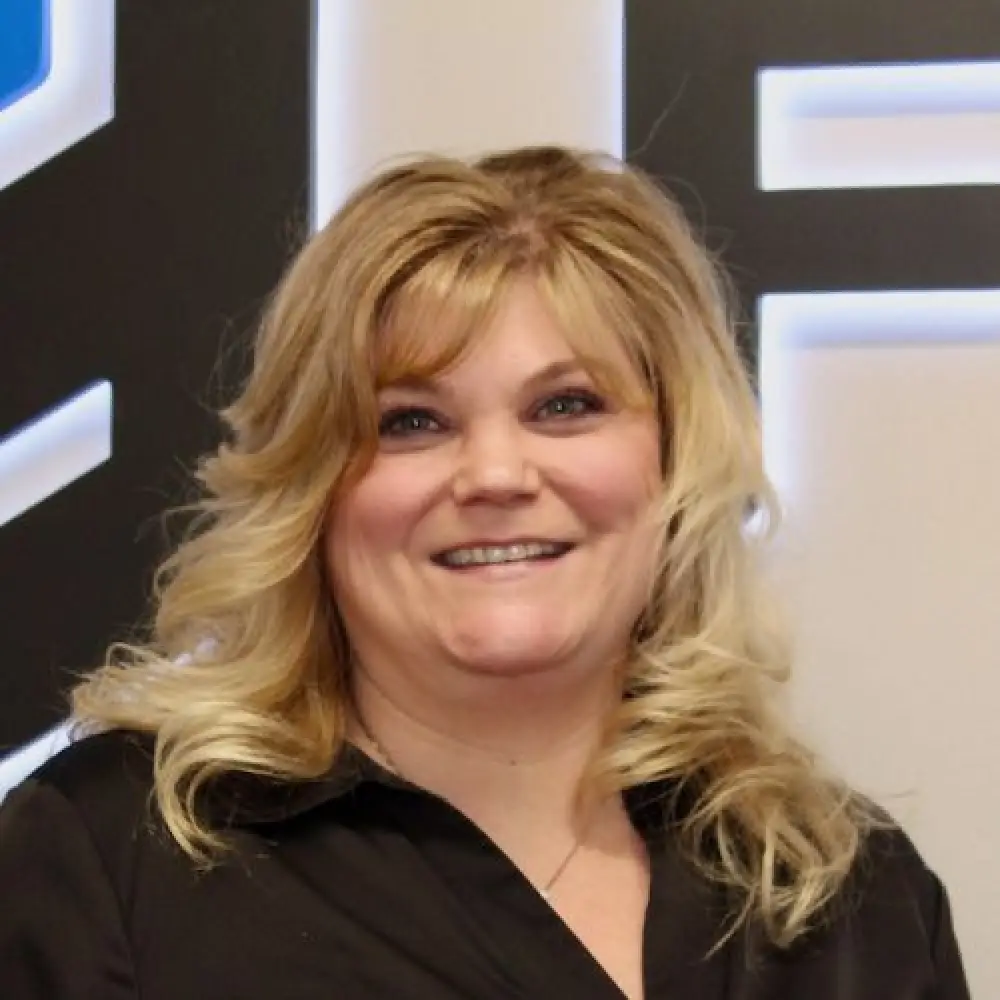 Smiling woman with wavy blonde hair in a black shirt, standing indoors.