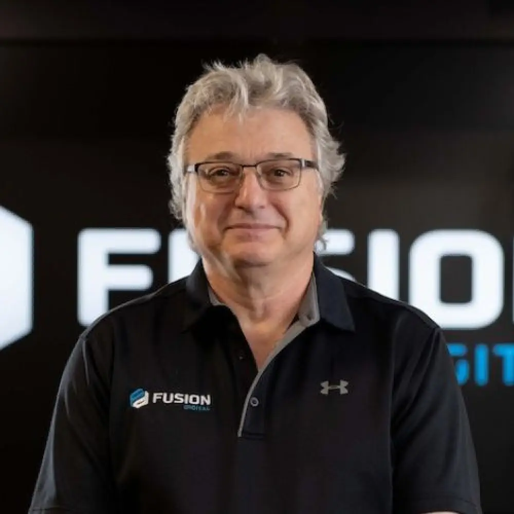 Older man with gray hair and glasses, wearing a Fusion logo polo, stands indoors smiling.