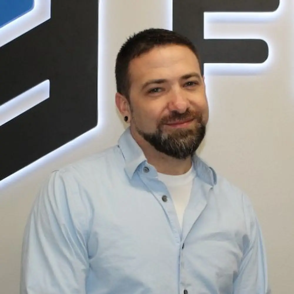 Man with short dark hair and beard, wearing a light blue shirt, standing by a lit wall sign.
