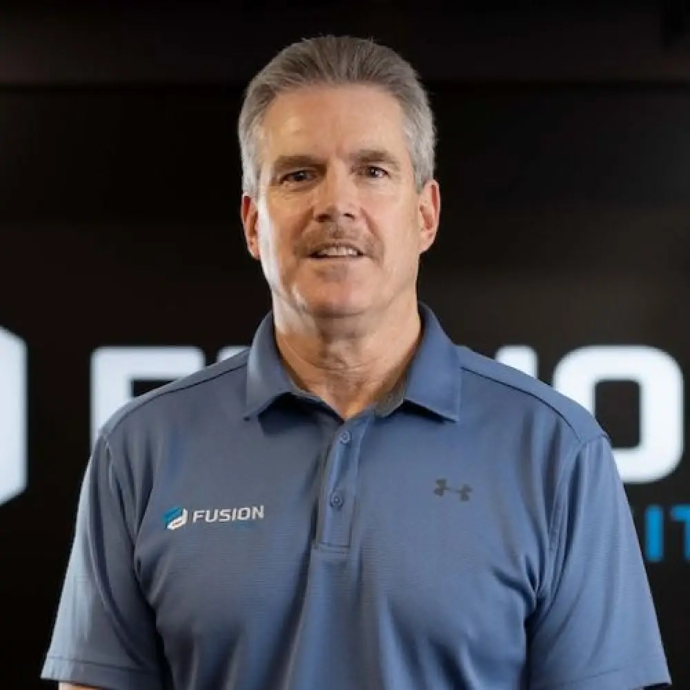 Man with gray hair and mustache wearing a blue Fusion polo shirt, standing indoors and smiling.
