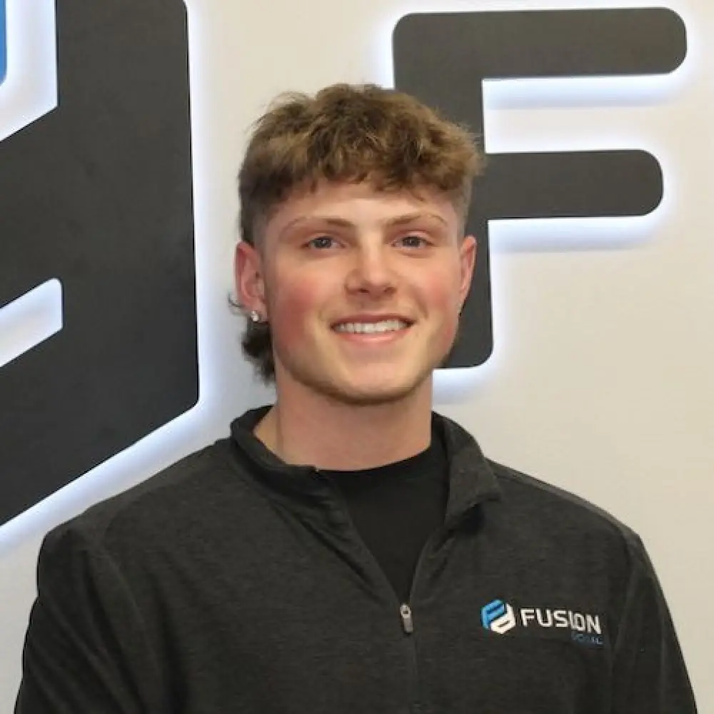 Young man with short, curly brown hair smiles, wearing a dark Fusion Social zip-up jacket.