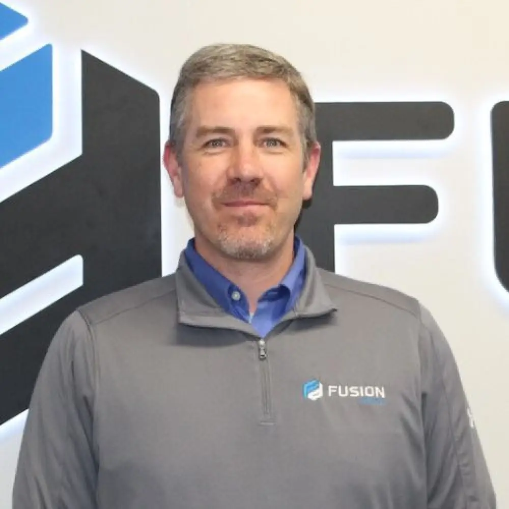 A man in a gray Fusion jacket stands in front of a wall with a Fusion logo and sign.