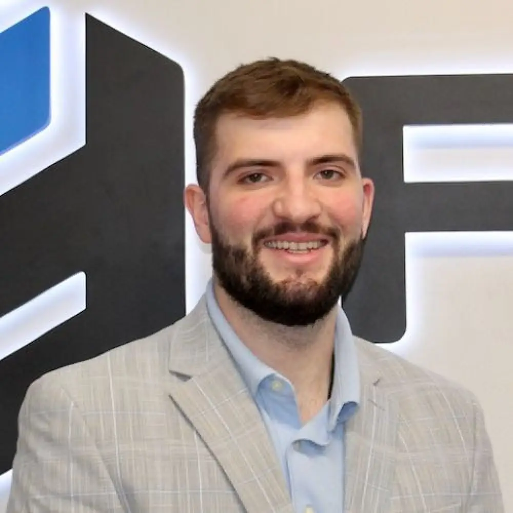 Smiling man in a light gray suit jacket stands in front of a large logo on the wall.