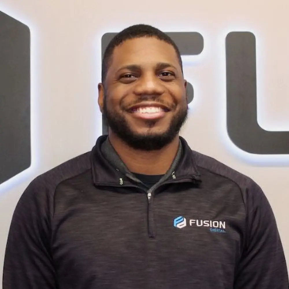 Smiling man in a black Fusion Digital shirt standing in front of a lighted wall logo.