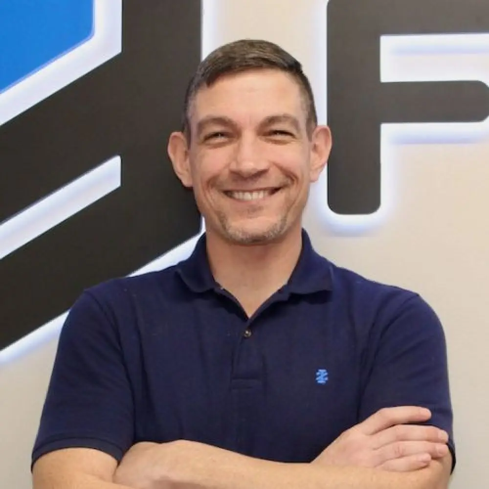 Smiling man in a navy polo shirt stands with arms crossed in front of a modern wall sign.