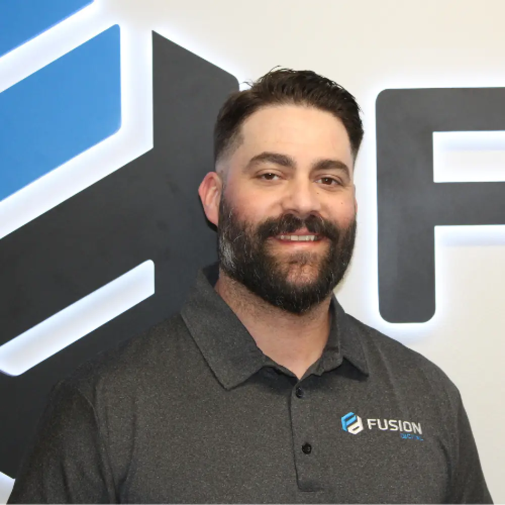 Man with a beard smiling, wearing a Fusion polo shirt, standing in front of a logo wall.
