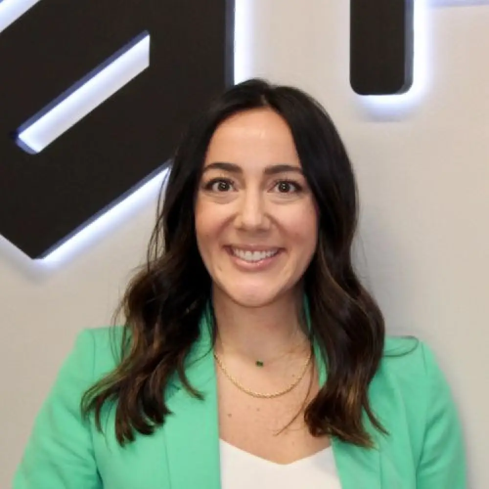 Smiling woman with long dark hair in a green blazer, standing indoors against a wall.