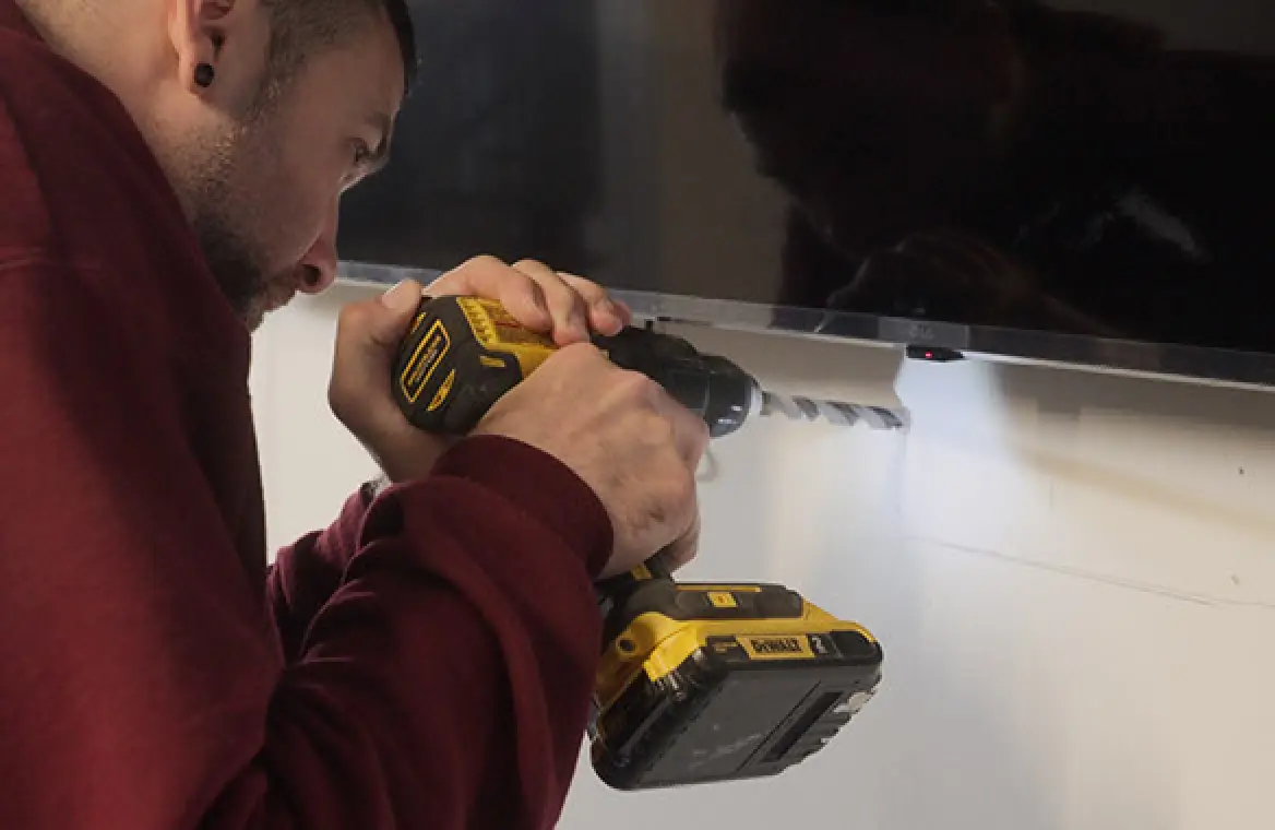 A man uses a power drill to make a hole in a wall beneath a mounted TV.