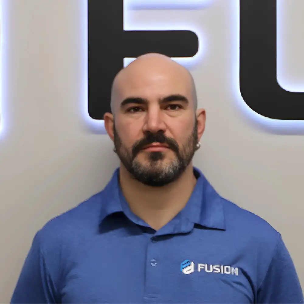 Bald man with a beard in a blue Fusion shirt stands against a backlit wall.
