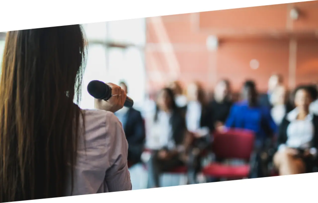 Person holding a microphone and speaking to an audience seated in a bright, modern room.