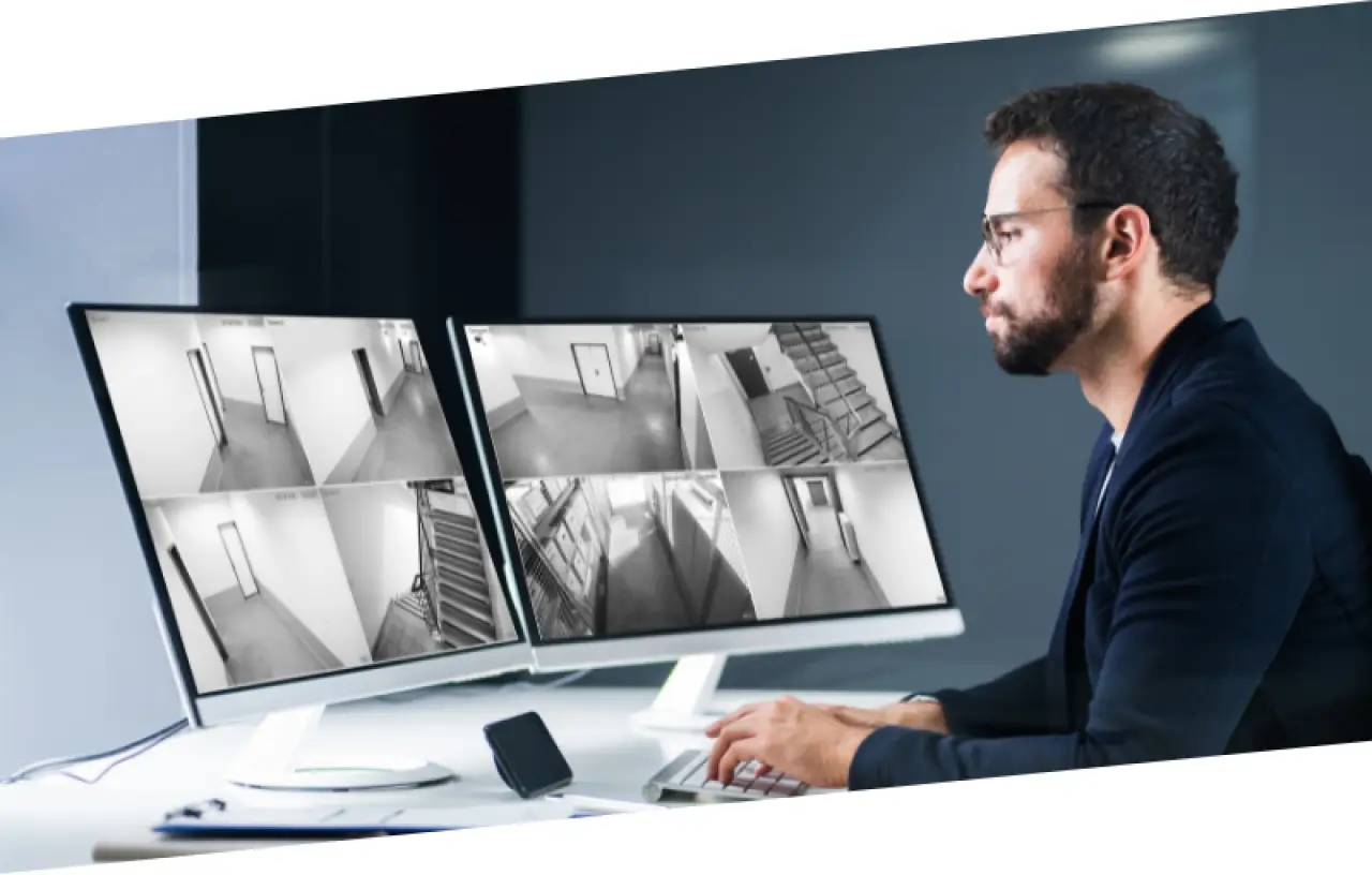 Man monitoring building security cameras on two computer screens in a modern office.