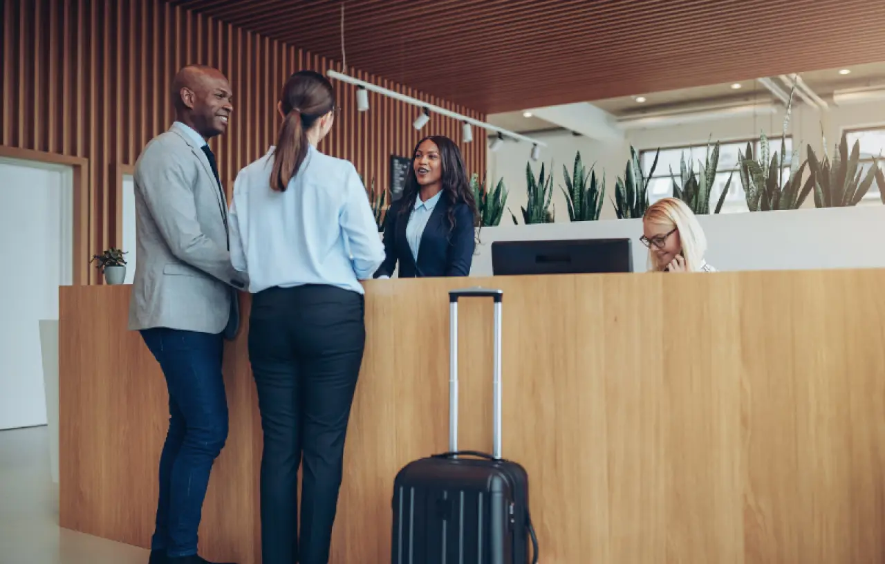 Two people check in at a hotel front desk, talking to a receptionist, with a suitcase nearby.