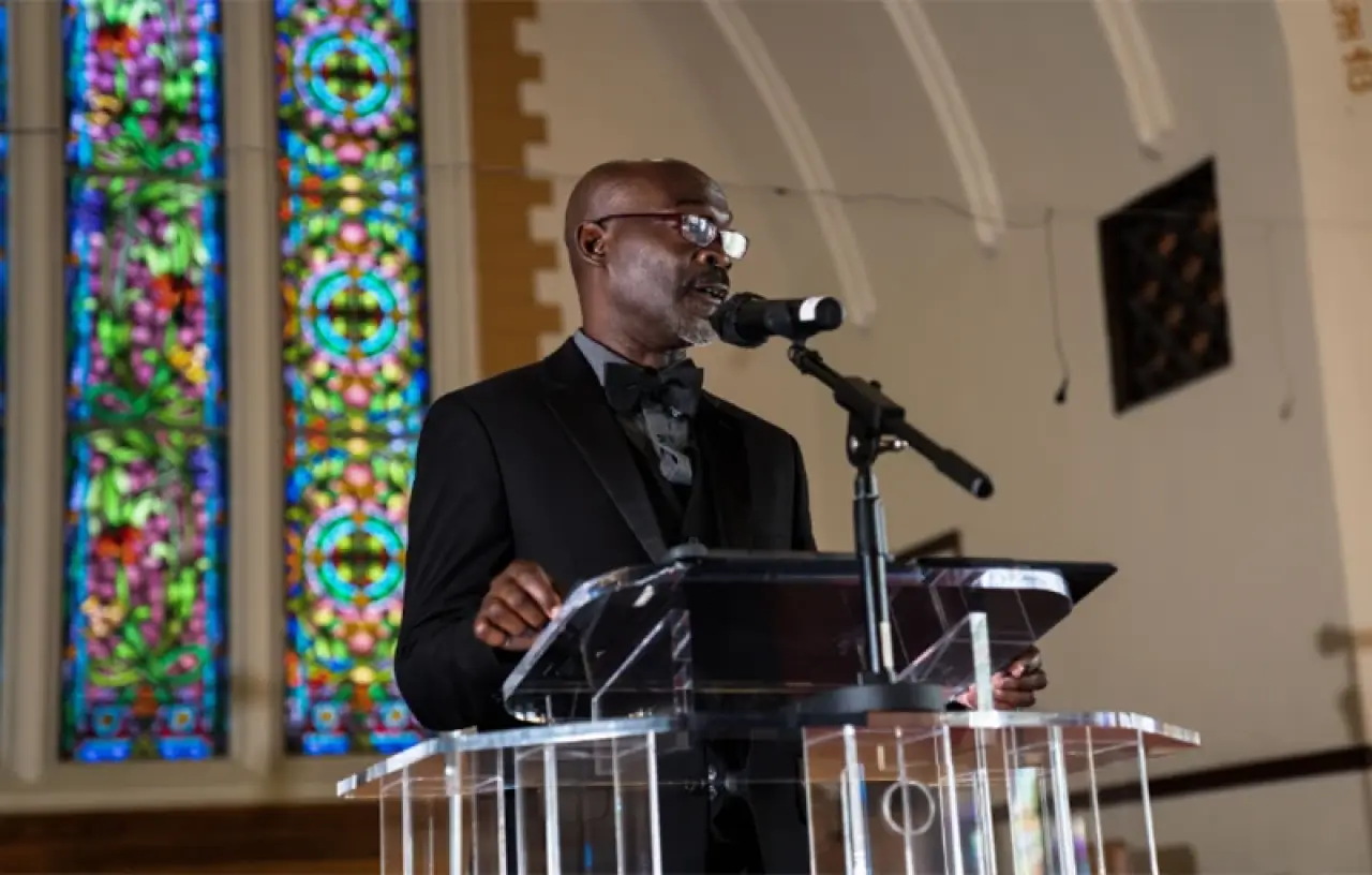 A man in a suit speaks at a podium in a church with stained glass windows behind him.
