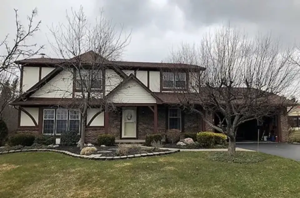 Two-story suburban house with brick and siding exterior, leafless trees, and a cloudy sky.