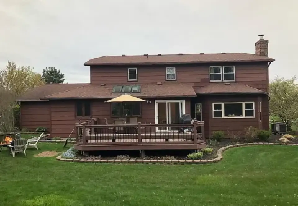 A two-story brown house with a wooden deck and patio furniture in a green backyard.