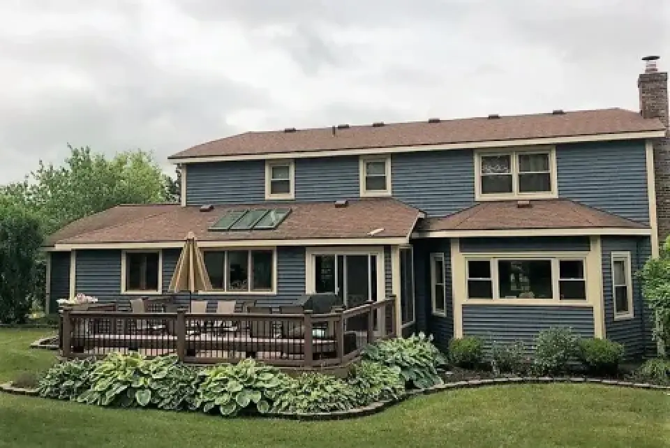 Two-story blue house with a brown roof, backyard deck, patio furniture, and lush greenery.