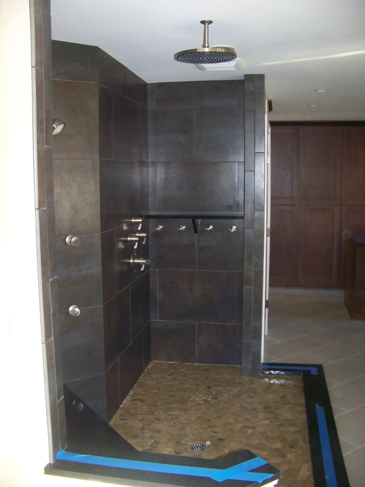 A modern shower with dark tiled walls, multiple showerheads, and a pebble floor. The area is partially finished.