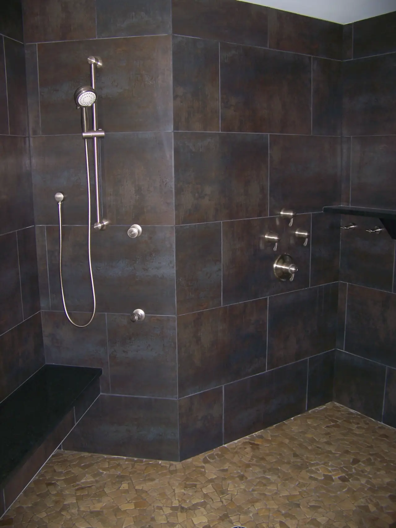A modern shower with dark tiled walls, a handheld showerhead, and a stone bench. The floor features mosaic tiles.