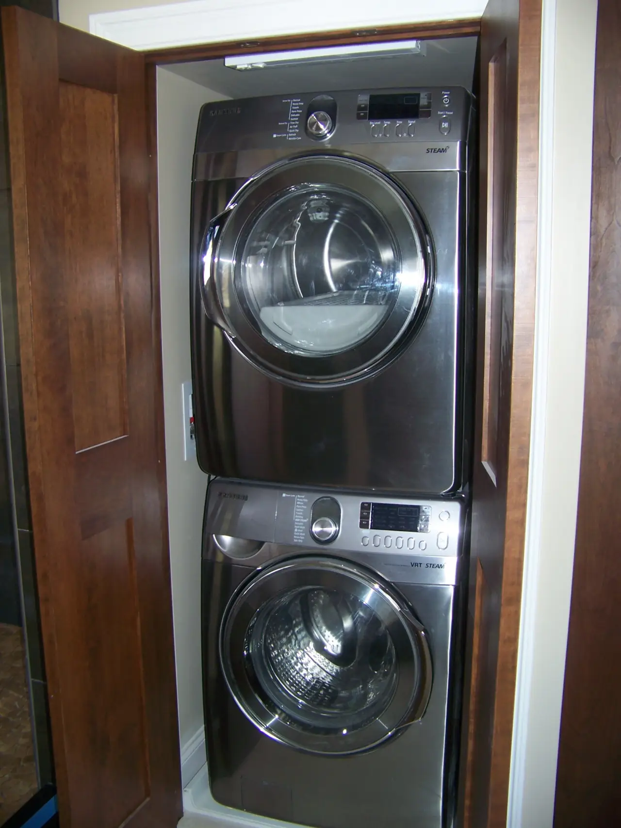 A stacked washer and dryer in a wooden cabinet, featuring a sleek metallic finish and control panels on the front.