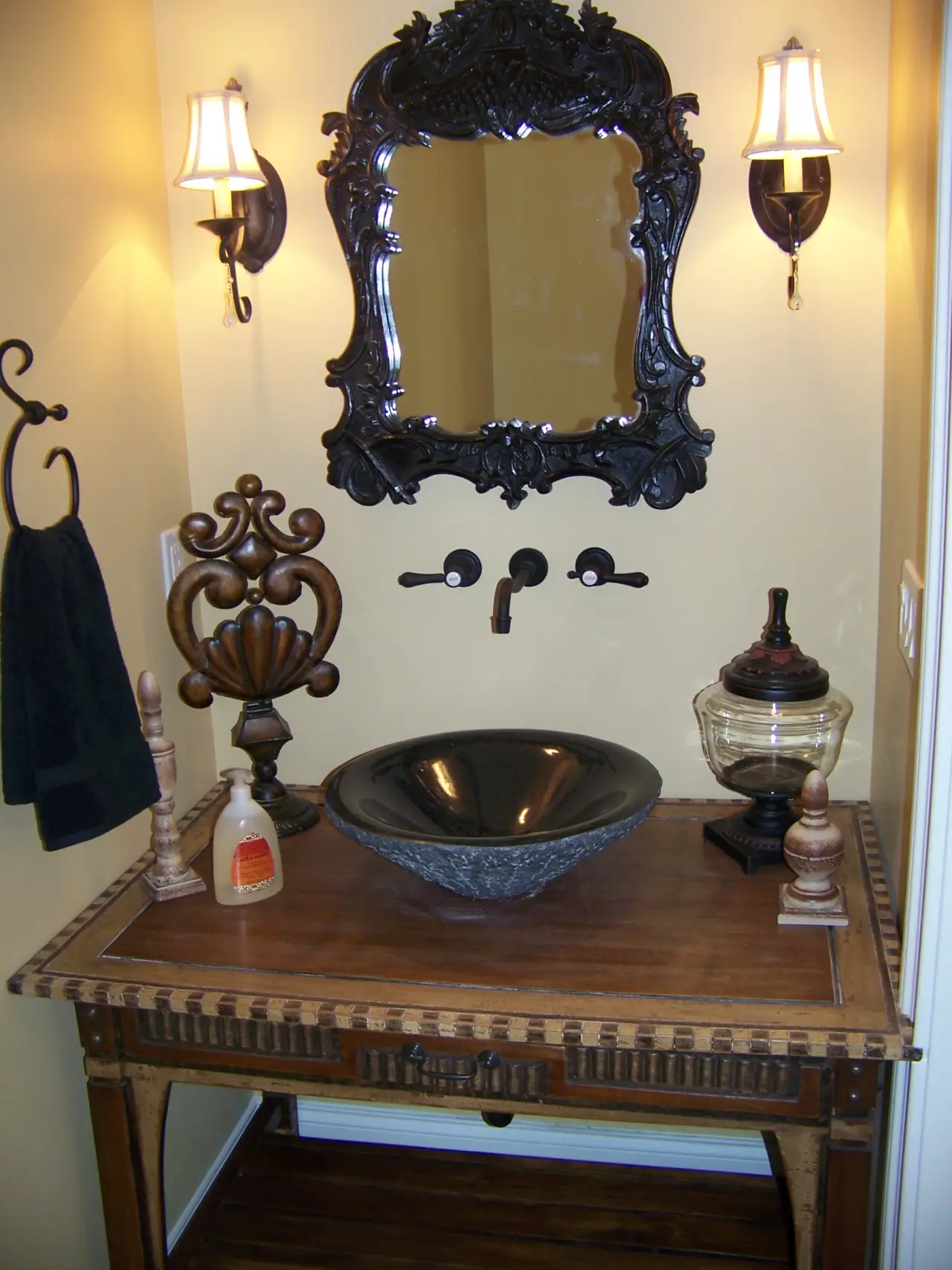 A vintage-style bathroom vanity with a black bowl sink, ornate mirror, wall sconces, and decorative accessories.