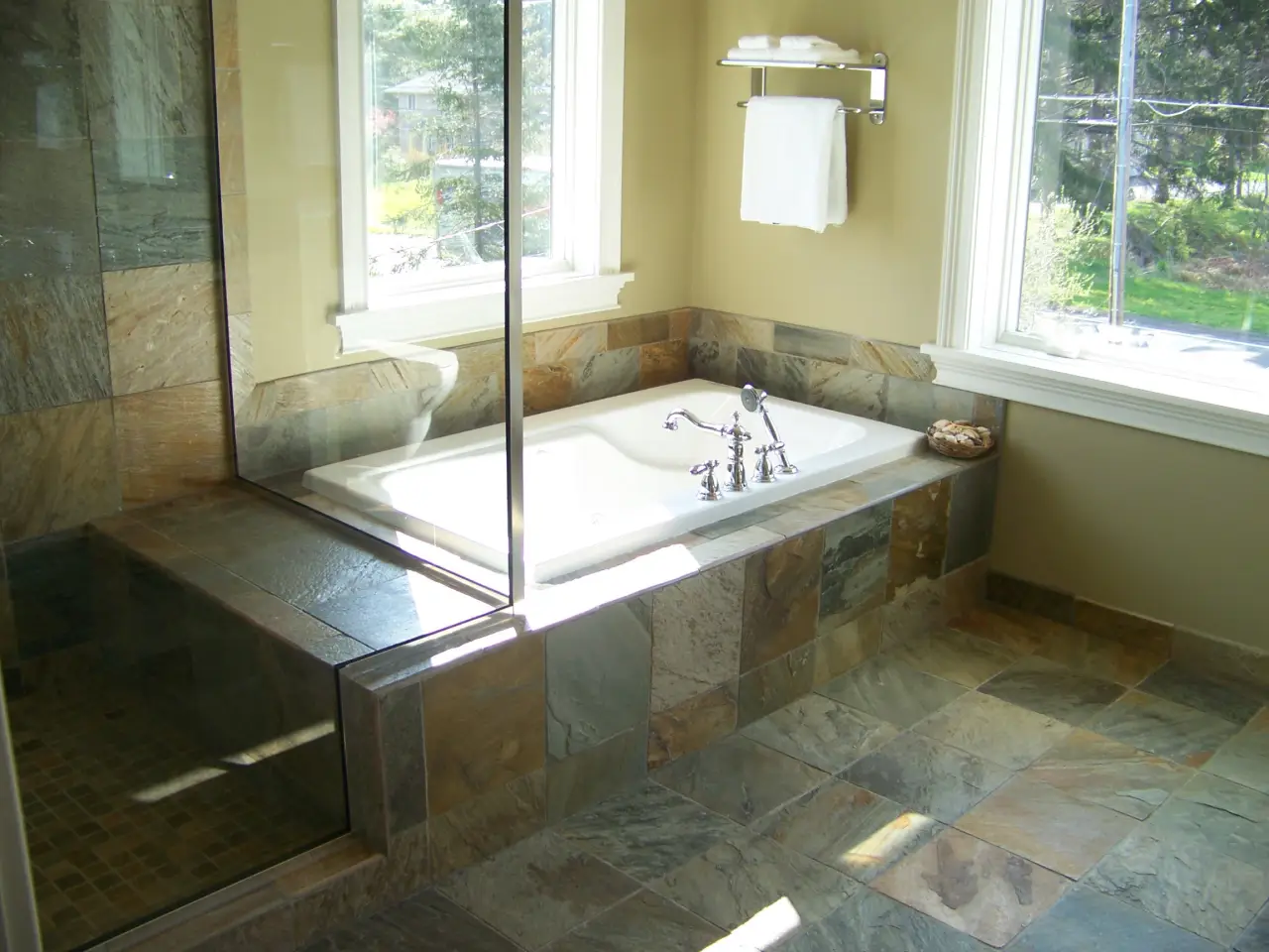 A modern bathroom featuring a glass shower, a large soaking tub, and slate tile flooring, with natural light from windows.