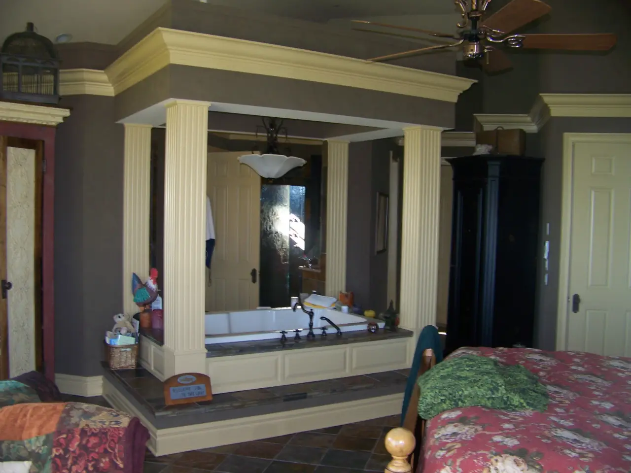 A spacious bathroom with a large tub framed by columns, a ceiling fan, and decorative elements, adjacent to a bedroom.