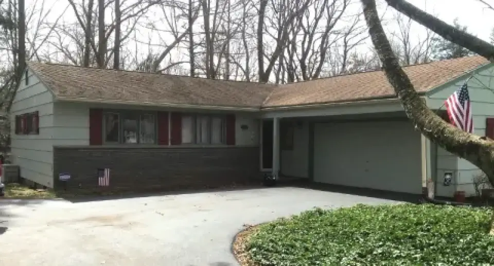 Single-story house with a two-car garage, American flag, and trees in the background.