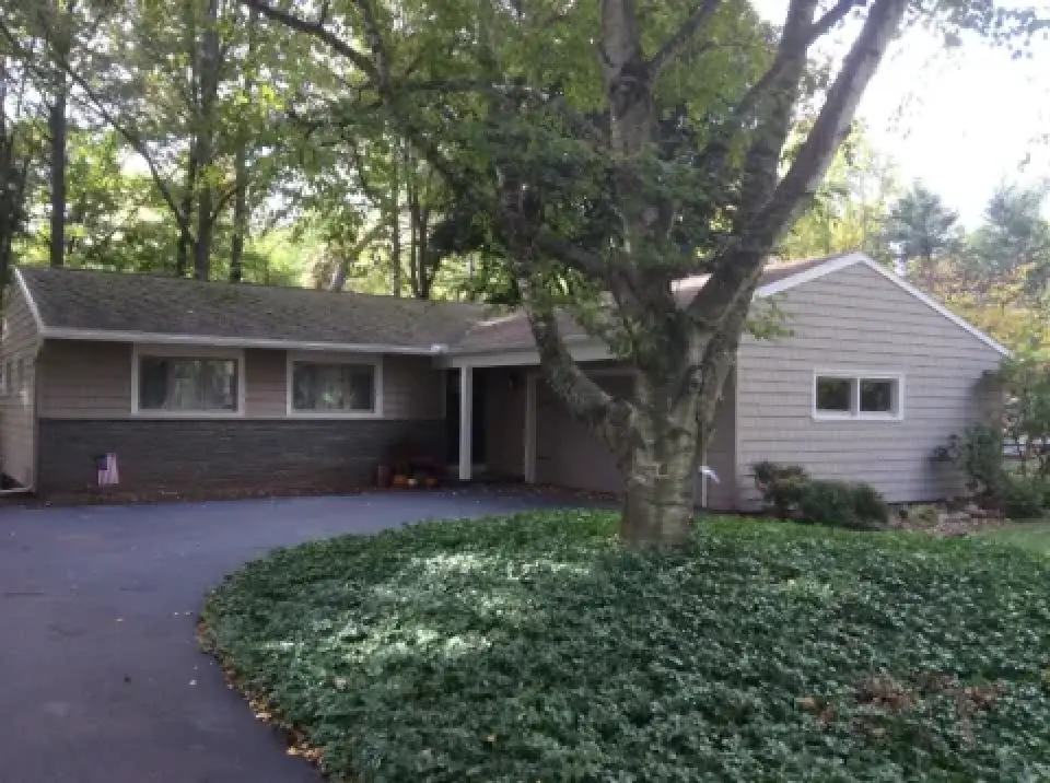Single-story house with gray siding, a large tree, and a driveway in front, surrounded by greenery.