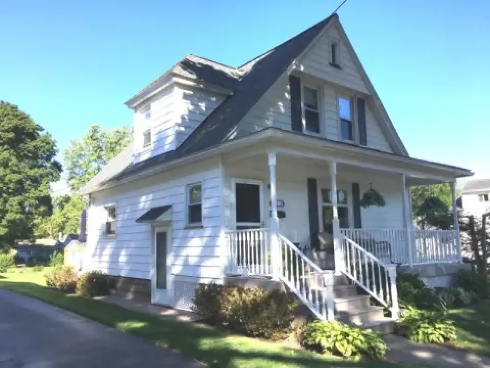 A white two-story house with a front porch, sloped roof, and green shrubs in the yard.