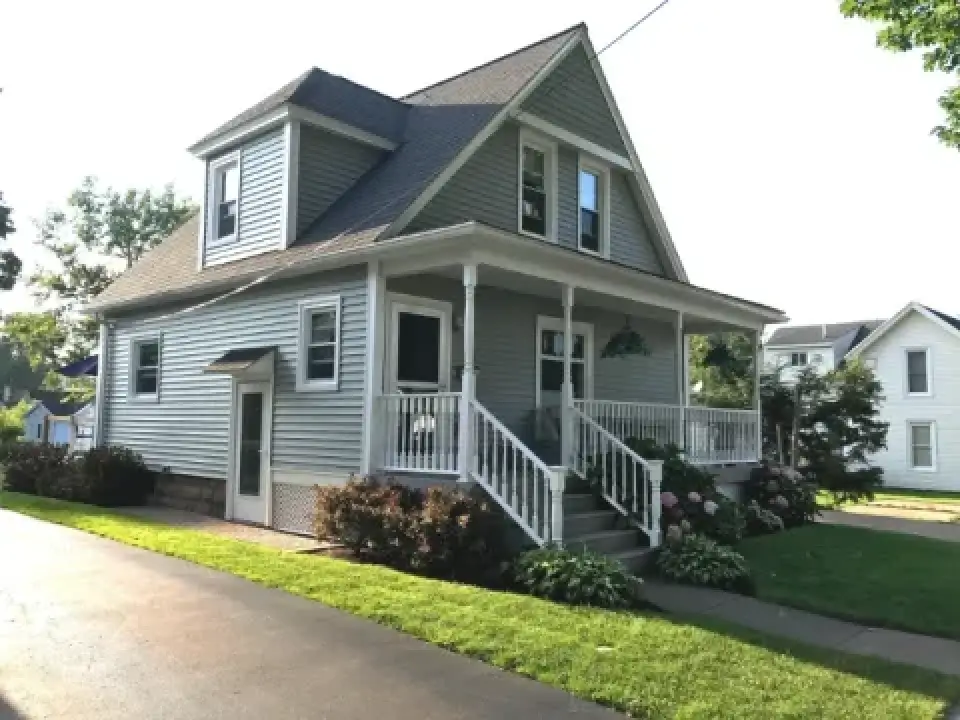 Light gray, two-story house with white trim, front porch, and a well-kept lawn on a sunny day.