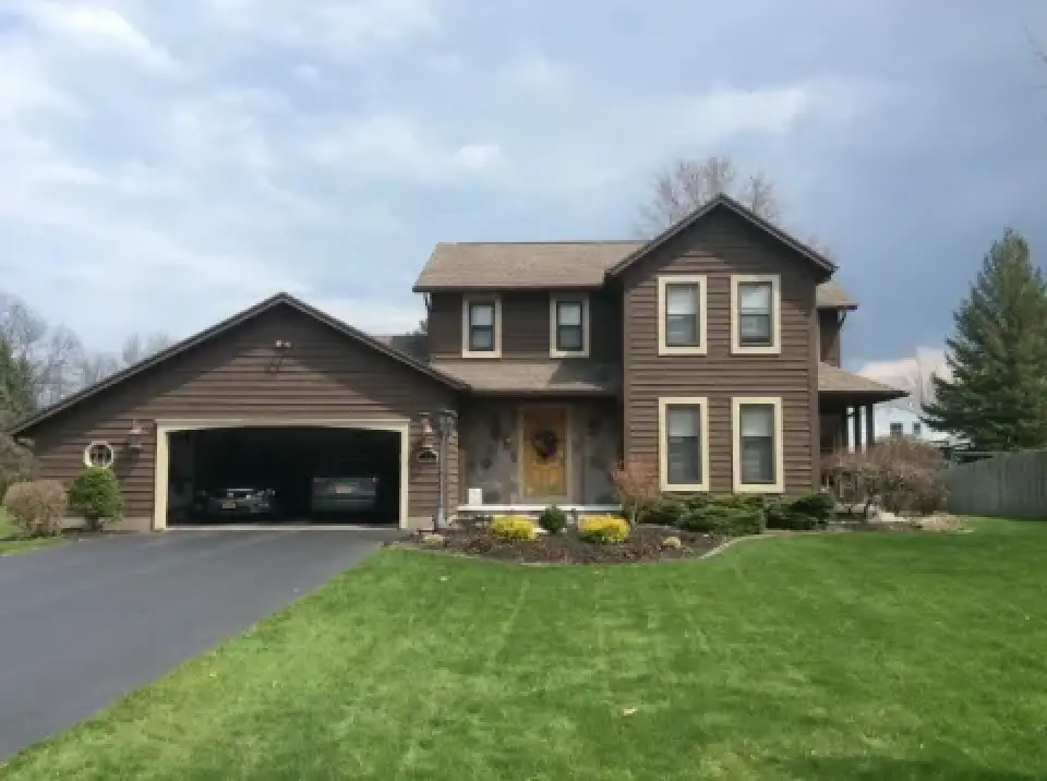 Two-story brown house with attached garage, green lawn, shrubs, and a paved driveway.