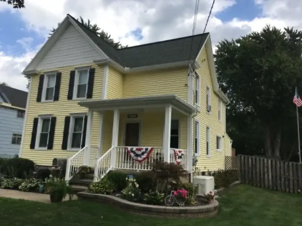 Yellow two-story house with white trim, porch, and patriotic bunting, surrounded by greenery.