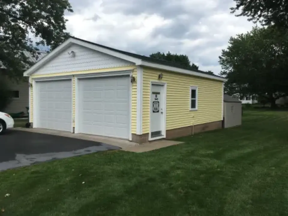 A yellow detached garage with two white doors and a white entry door sits on a green lawn.