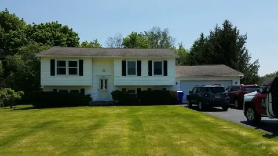 A white split-level house with black shutters, front lawn, and cars parked in the driveway.