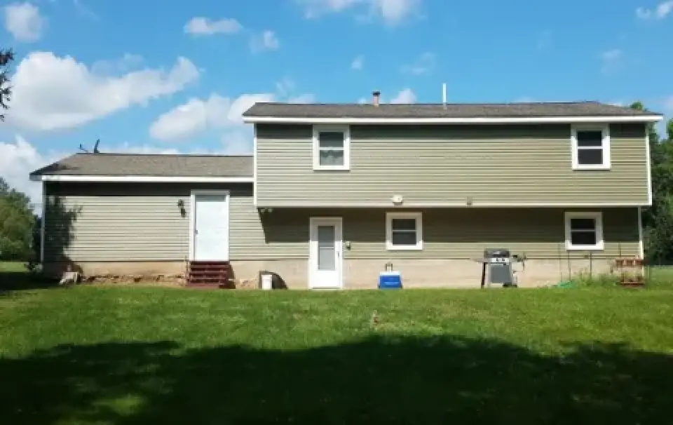 Two-story tan house with white trim, grassy yard, grill, and blue sky with scattered clouds.