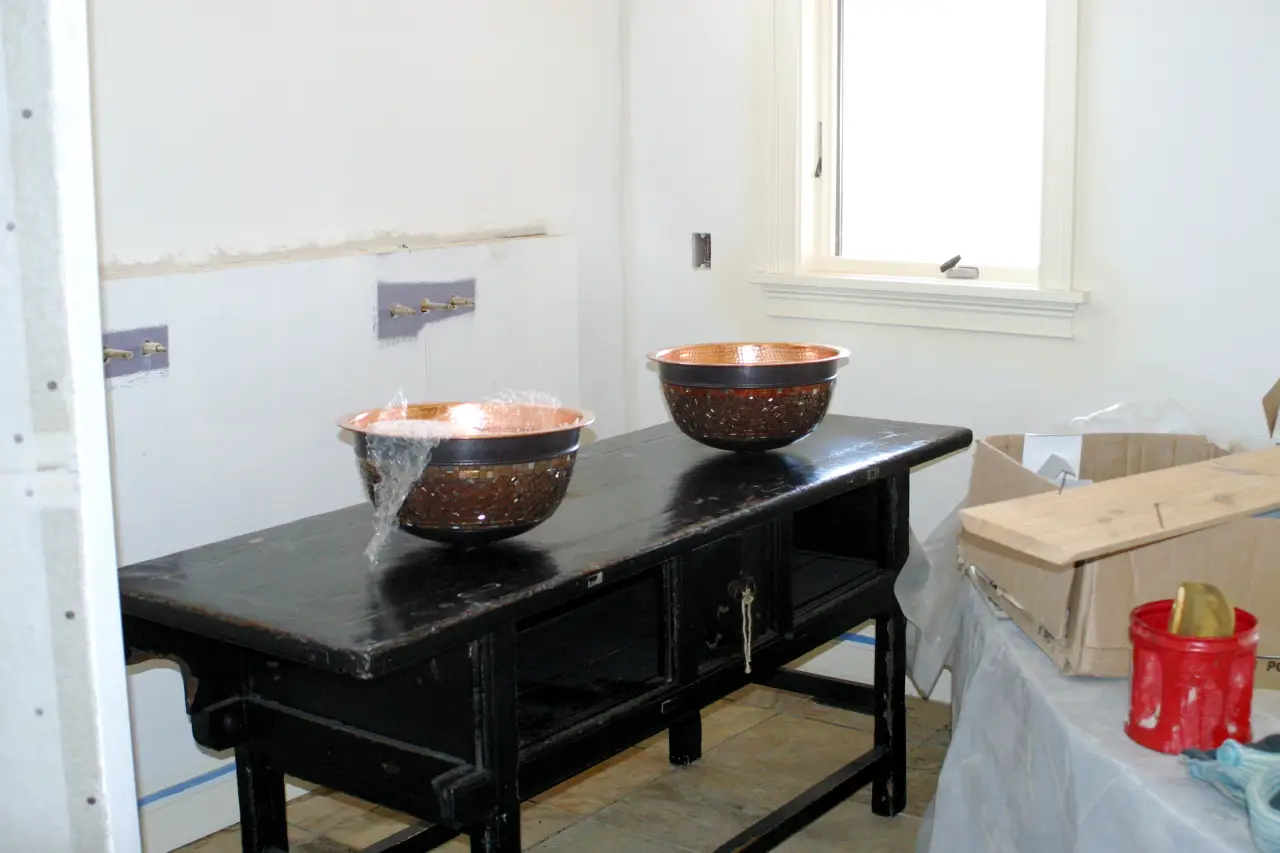A black table with two copper bowls on top, set against a bare wall with plumbing fixtures. A box and a red bucket are nearby.