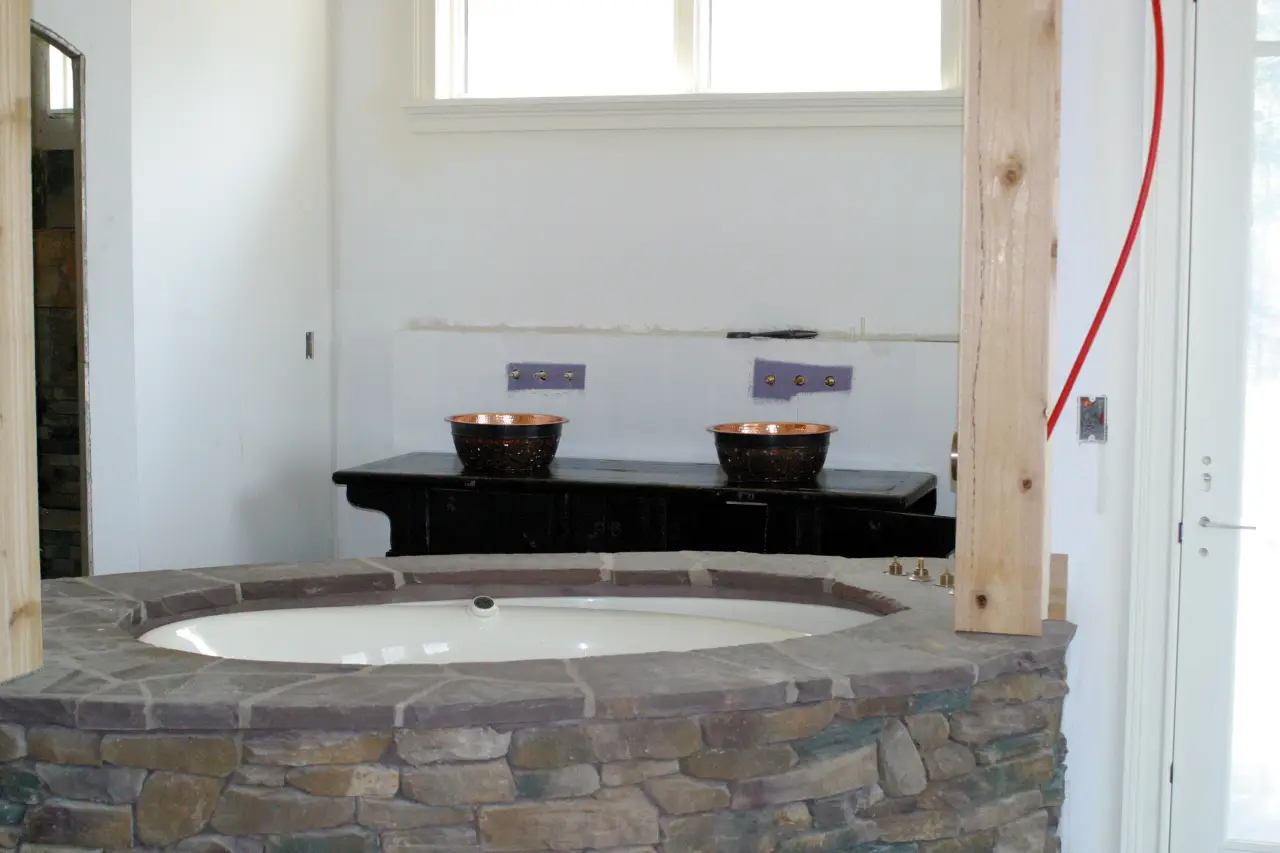 A bathroom featuring a stone tub, two copper sinks on a dark wooden counter, and large windows for natural light.