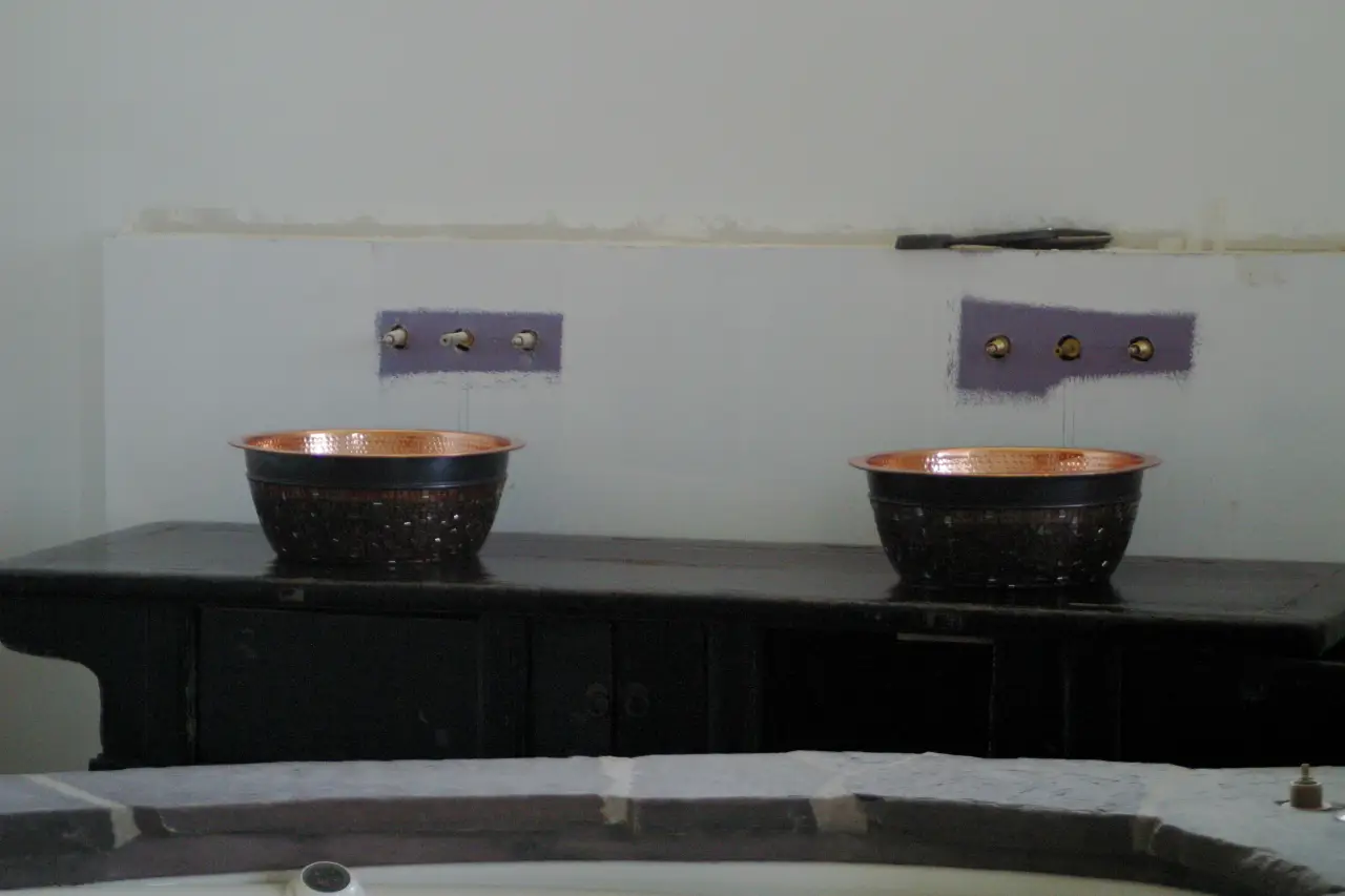 Two copper bowls sit on a dark wooden surface, with wall fixtures above them and unfinished wall sections in the background.