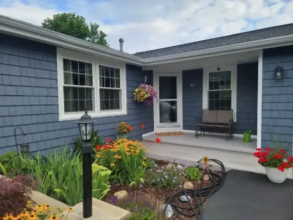 Blue house with a small porch, flowers, a bench, and a hanging basket on a sunny day.